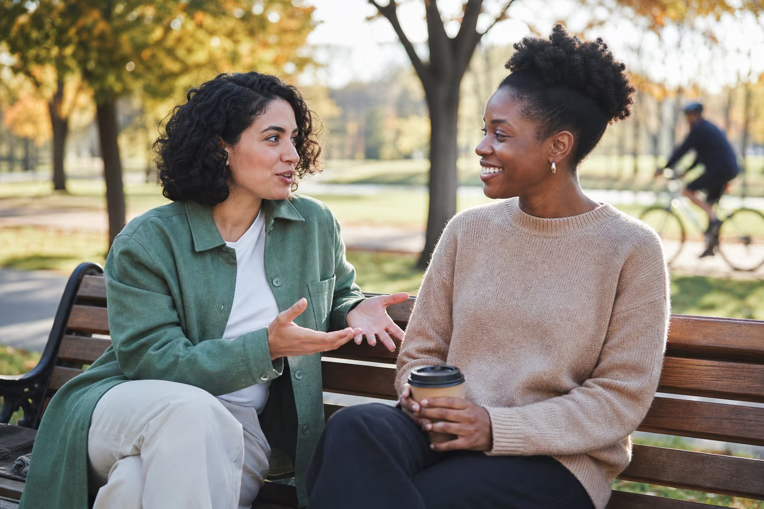 Two friends sitting on a park bench in the sun, one talking with their hands, the other listening with a coffee cup