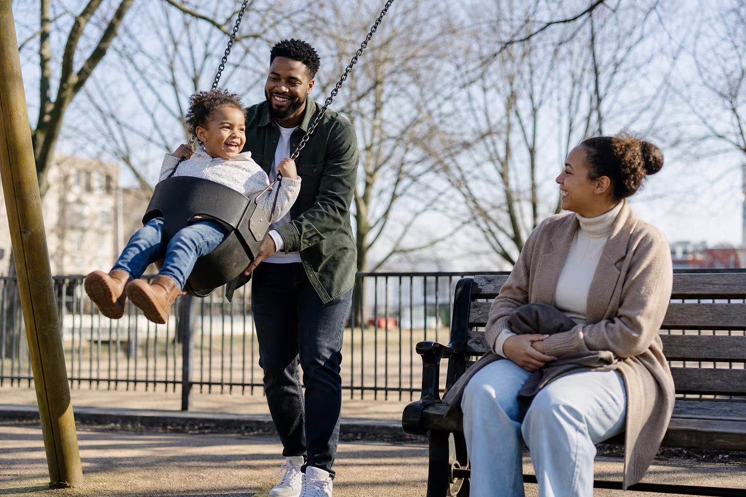 A parent and a young child on a swing in a park while another adult watches from a bench nearby