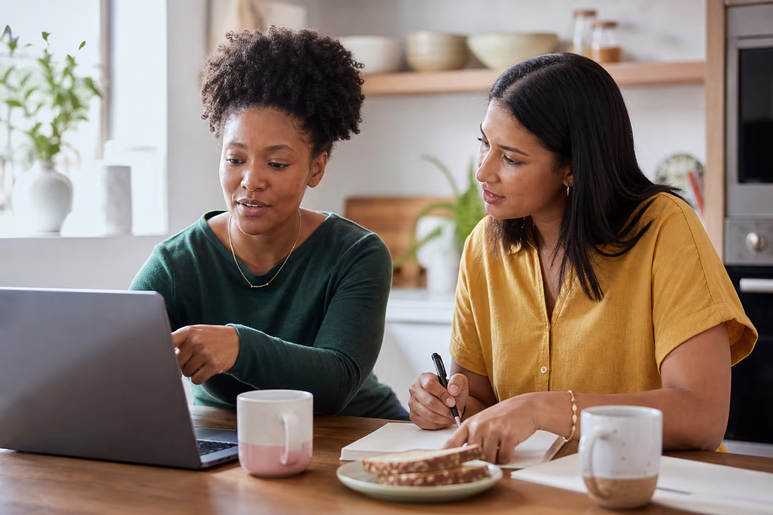 Two people at a kitchen table comparing notes on a laptop over morning coffee