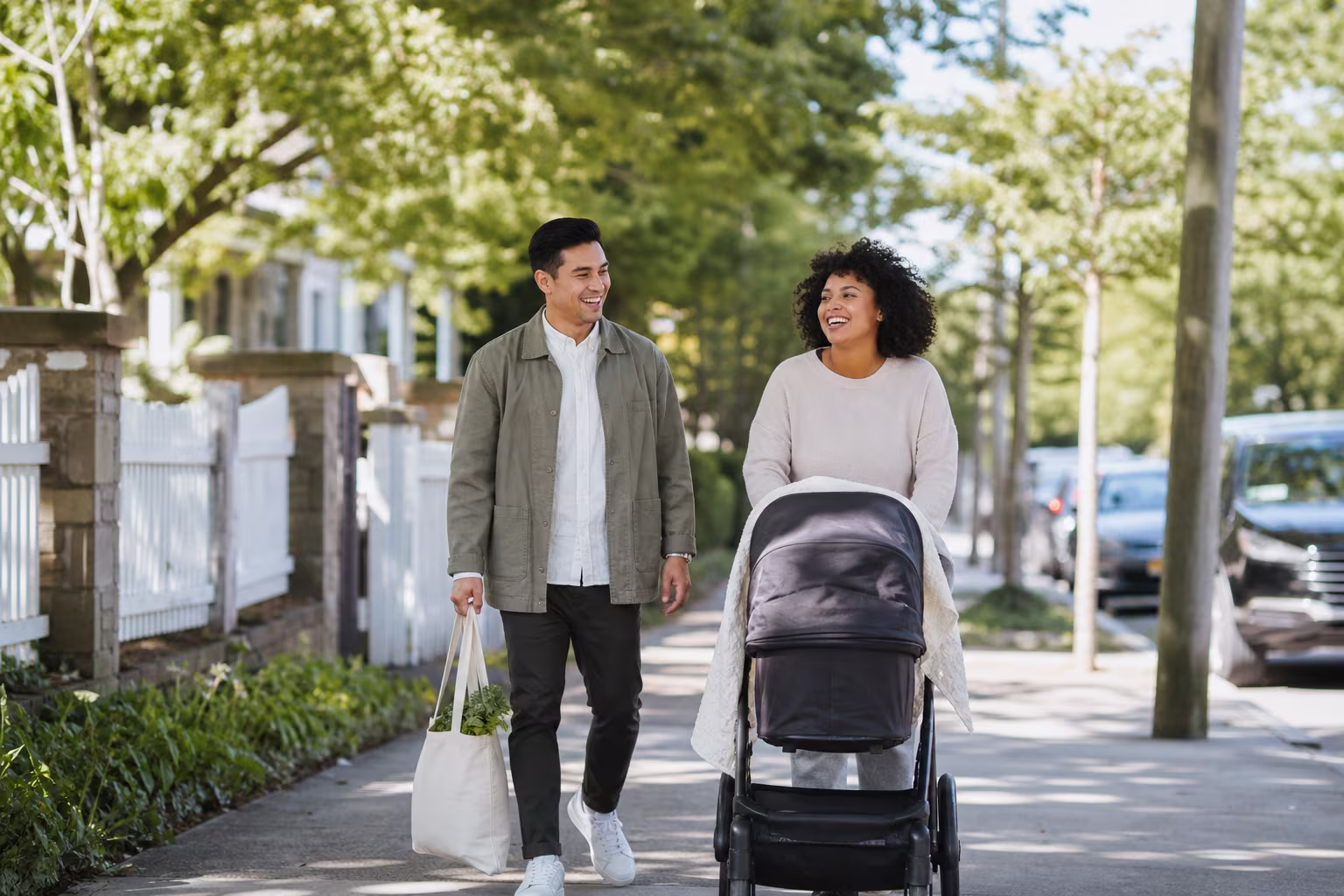 A parent pushing a pram along a tree lined residential street on a bright morning with another adult walking alongside