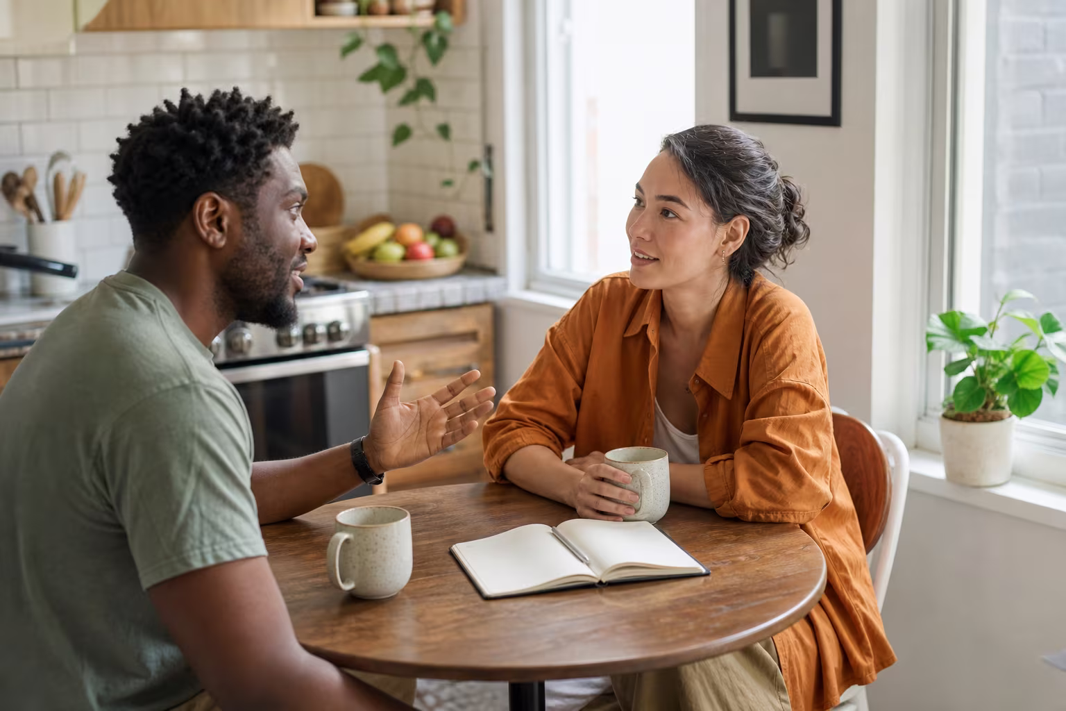 Two people talking over coffee at a kitchen table with a notebook between them