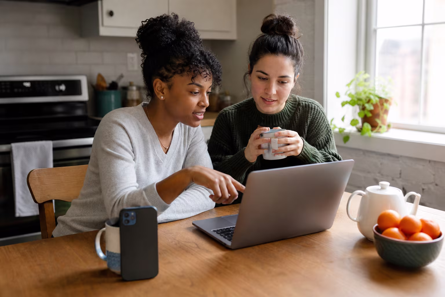 Two friends at a kitchen table comparing donor profiles on a laptop and a phone over mugs of tea