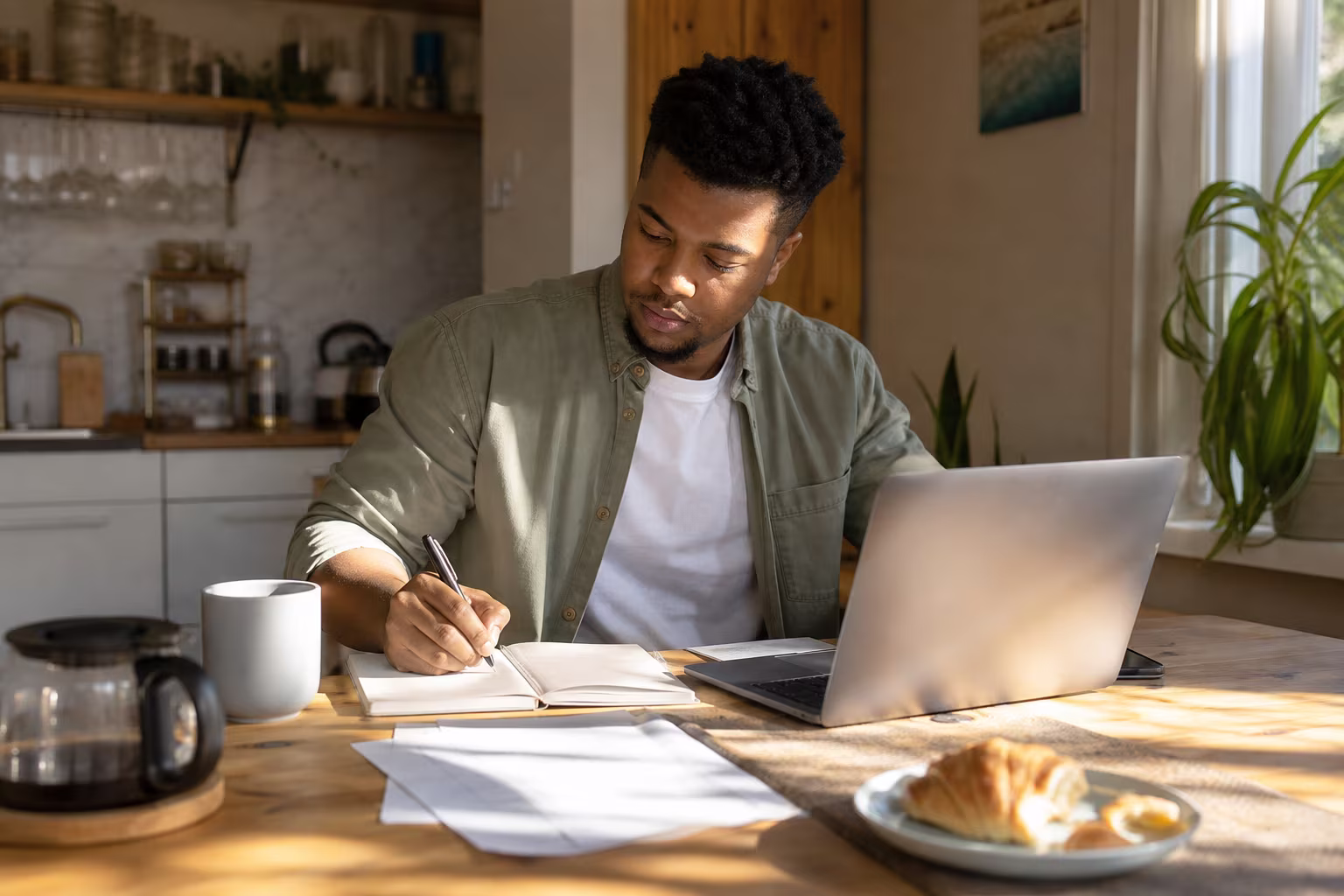 A person at a kitchen table with a laptop and a notebook, working through household costs on a weekend morning