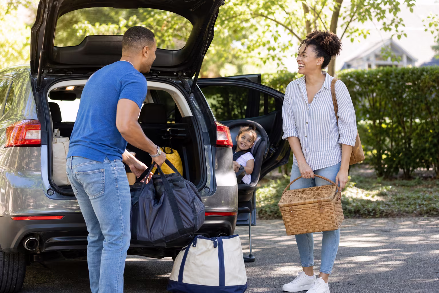 A family loading bags into a car in a driveway with a toddler in a car seat on a bright afternoon