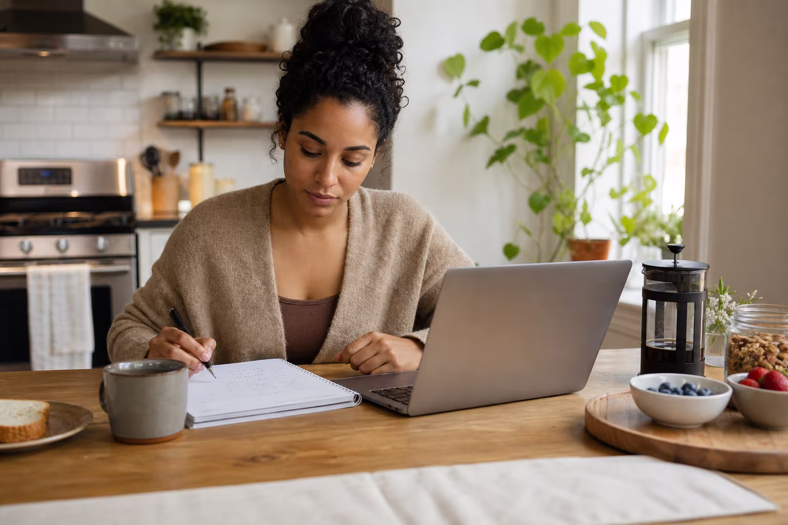A woman at a kitchen table with a laptop, a notebook and a mug, working through numbers on a quiet morning