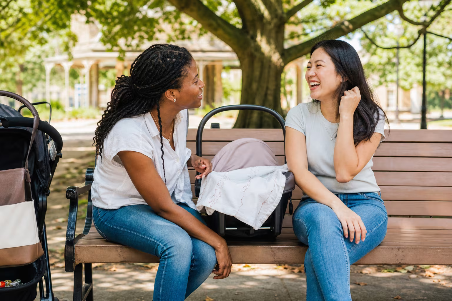Two friends sitting on a park bench with a baby carrier between them, talking on a bright afternoon