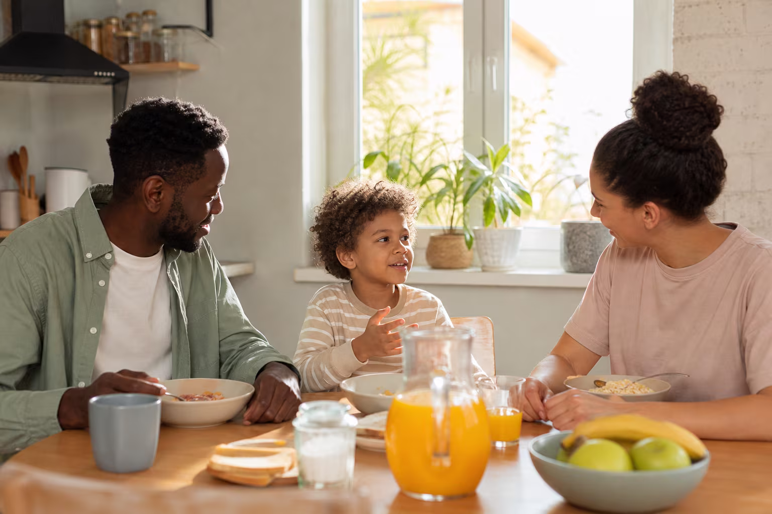 Two parents and a child eating breakfast together in a bright kitchen, talking easily