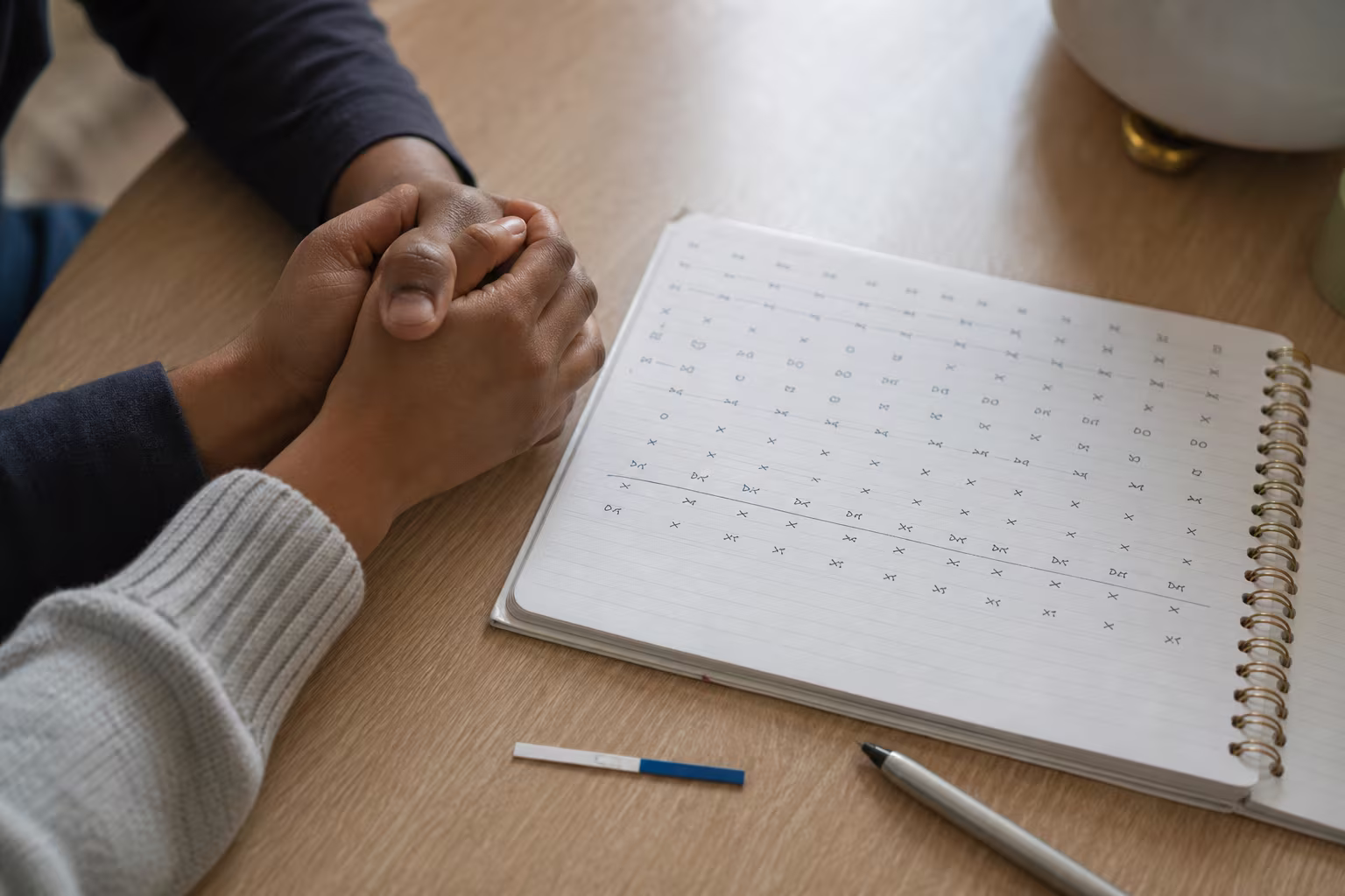 Hands beside a notebook of handwritten cycle dates and an ovulation test strip on a desk